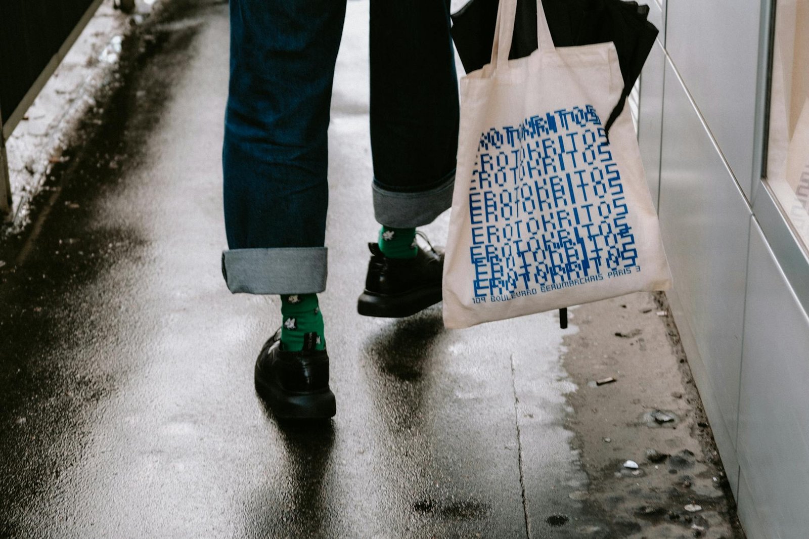 Casual urban stroll with tote bag, denim jeans, and black shoes on a wet city street.